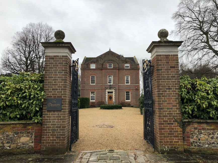 Photograph of a large, three-storey brick house viewed through an ornate black metal gate supported by two tall brick pillars topped with stone spheres. The gate opens onto a gravel driveway leading directly to the house, which has a central wooden door framed by stone detailing, symmetrical windows on each side, and additional windows on the upper floors. Flanking the driveway are well-maintained shrubs and greenery, with leafless trees visible in the background under an overcast sky. The scene suggests a quiet residential property, possibly undergoing a home relocation process, with the area prepared for furniture transport and moving logistics handled by Man with Van Lamorbey.
