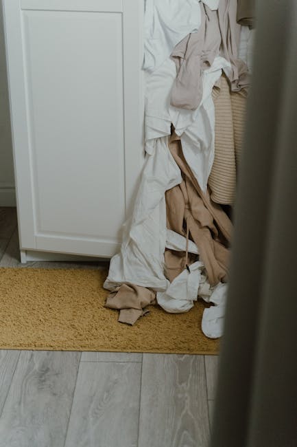 Inside a residential property, a pile of dismantled and crumpled fabric and packaging materials, including cardboard, paper wrapping, and cloth coverings, is seen on the floor next to a white wardrobe or cabinet. The materials are in various shades of beige, brown, and white, with some sections draped over the furniture. The scene suggests a home relocation or packing process, with the fabric and packaging likely used to protect and wrap furniture and household items. The box or wardrobe is positioned against a wall, with a yellowish mat on the light-colored wooden floor beneath the objects. The lighting appears natural, possibly from nearby windows, and the overall setting indicates an ongoing packing or loading activity facilitated by a professional removals service, such as Man with Van Lamorbey.
