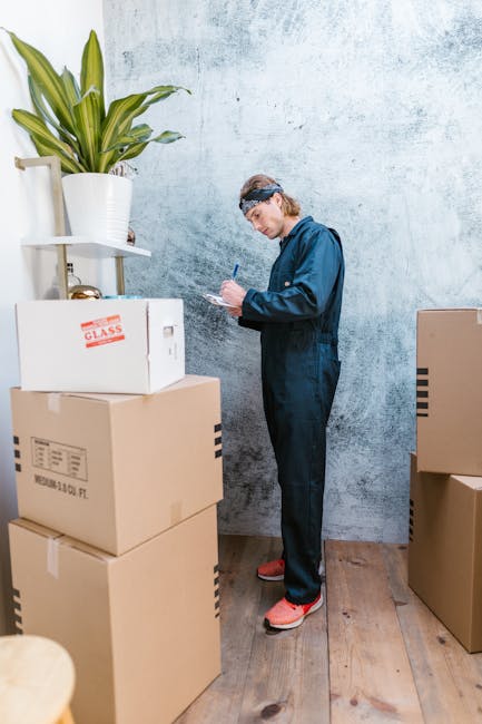 A man wearing a navy blue work jumpsuit and a protective headband is standing indoors against a textured grey wall, surrounded by several packed cardboard boxes of various sizes, some marked with labels like 'glass' and barcode stickers. The boxes are stacked on a wooden floor, with one placed on a small white shelf in the corner supporting a potted plant with long green leaves. The man is holding a pen and a clipboard, appearing to take notes or check inventory as part of a home relocation or packing process. The scene includes natural lighting, highlighting the moving environment typical of a house removals service, with the setting indicative of an ongoing packing and moving operation associated with services offered by Man with Van Lamorbey.