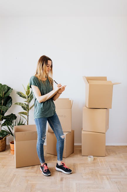 A young woman with blonde hair, dressed in a casual green T-shirt, ripped blue jeans, and pink and black sneakers, is standing indoors on a wooden floor, taking notes on a small notepad. Behind her, there are several unpacked cardboard moving boxes, some stacked one on top of the other, with one open box revealing its interior. To her left, a large green potted plant with broad leaves is situated against a plain white wall. The scene suggests she is preparing for a home relocation or move, with the boxes indicating packing and moving logistics, and the interior setting representing a typical moving environment supported by a professional removals service like Man with Van Lamorbey.