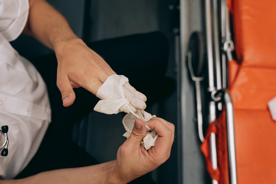Close-up of a person wearing a white uniform wrapping a piece of white cloth around their gloved hands during a furniture packing or loading process at a house removal. The individual is seated at a workbench or within a vehicle, with orange packing blankets, plastic wrapping, and packing tools visible nearby. In the background, there are metallic tools and equipment used for moving, such as straps and trolleys, indicating a professional furniture transport and home relocation setting. The scene highlights the careful packing and handling involved in prompt, professional same-day removals in Lamorbey, executed by Man with Van Lamorbey, with an emphasis on secure loading procedures and the use of protective materials to facilitate efficient house moving and furniture transport.