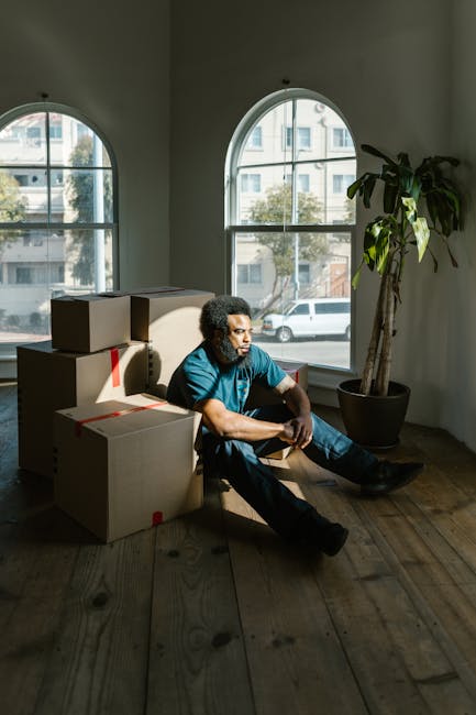 A man with dark curly hair and medium skin tone sitting on a wooden floor inside a room with large arched windows that let in natural light. He is wearing a blue shirt and dark pants, with a neutral e