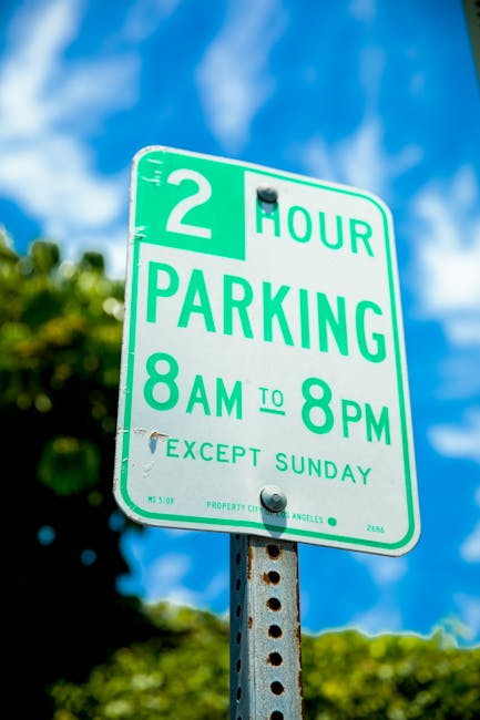 A green and white parking restriction sign mounted on a metal post outdoors against a blue sky with some clouds and blurred green foliage in the background. The sign indicates a two-hour parking limit from 8 am to 8 pm, except on Sundays, with the text '2 HOUR PARKING 8AM TO 8PM EXCEPT SUNDAY' clearly visible. The sign is positioned on a paved surface, and its purpose is to communicate parking rules in Lamorbey, which is relevant for home relocations or moving logistics in the area, as managed by Man with Van Lamorbey. The image captures the sign's details accurately, highlighting its role in local parking regulations that may impact furniture transport and house moving activities.
