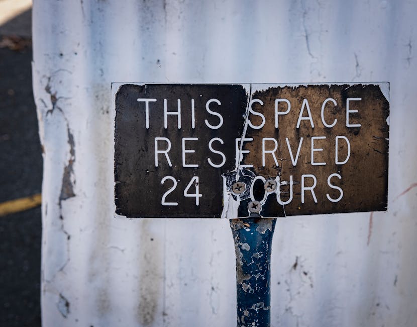 Close-up of a weathered metal parking restriction sign mounted on a blue-painted pole, attached to a white wall with visible peeling paint and cracks. The sign displays the message 'THIS SPACE RESERVED 24 HOURS' in white uppercase letters on a black background, with some rust and damage visible around the edges. The pole and sign are situated outdoors, and the photograph emphasizes the aged, textured surface of the sign amid an urban environment. This image is relevant to house removals and logistics, as parking regulations can impact the planning of home relocation and furniture transport services, which are offered by Man with Van Lamorbey.