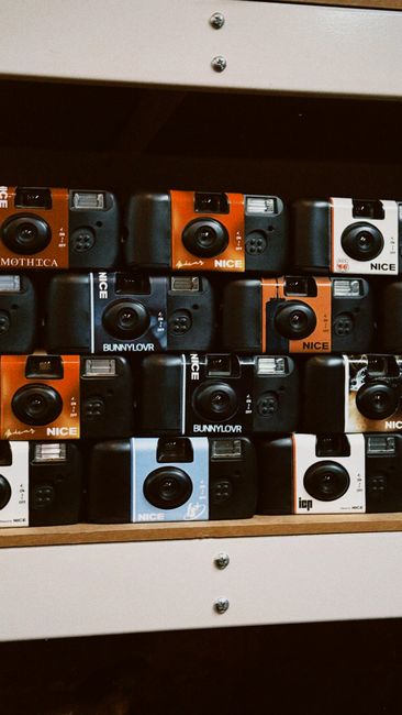 A collection of compact instant cameras arranged on a wooden shelf inside a property, with various models including orange, black, gray, and white units, some branded 'NICE' or 'BUNNYLOVR'. The shelf is mounted on a wall, with two visible screws securing it, and the cameras are organized in rows, some facing forward, others slightly angled. The area is well-lit, and the arrangement suggests preparation for a home relocation or packing process involving camera equipment, which is part of a wider move managed by Man with Van Lamorbey. The scene reflects the careful handling of small electronic items during furniture transport, characteristic of professional removals and moving services.