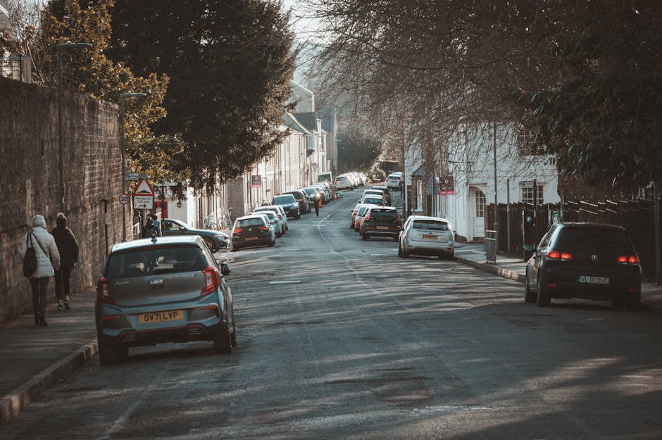 A narrow residential street with a slight incline during daylight, lined with parked cars on both sides including a silver hatchback in the foreground with the registration OV71 LVP and a black vehicle further along. The street features a brick wall on the left and trees with sparse foliage casting shadows on the pavement and buildings. Sidewalk pedestrians are visible, and the scene is lit by natural sunlight, highlighting the urban environment typical of house removals and home relocation logistics. The image supports services such as furniture transport and moving logistics, showcasing the challenge of navigating narrow street access for moving vehicles, as handled by companies like Man with Van Lamorbey.