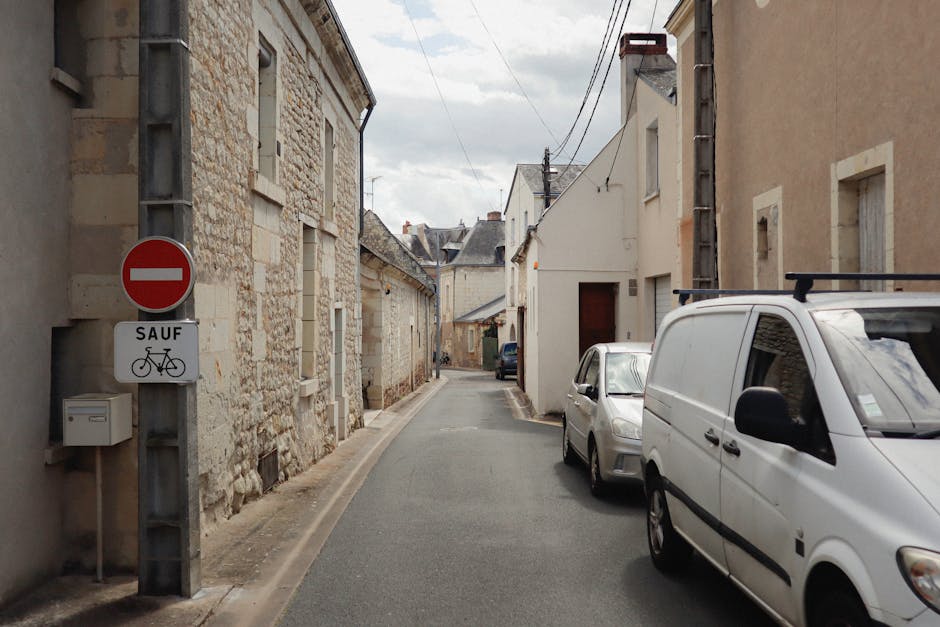 A narrow residential street with closely spaced stone and brick buildings on both sides, some with window shutters and small doors. Parked vehicles, including a white van and a silver hatchback, are positioned along the pavement, with the van appearing to be in the process of loading or unloading. Overhead, utility wires run parallel to the street, and there is a street sign indicating no entry except for bicycles, mounted on a metal pole on the left side. The pavement is narrow and the scene suggests a tight clearance suitable for house removals and furniture transport in confined urban environments. Man with Van Lamorbey's moving services might be involved in this home relocation, handling the logistics of navigating narrow streets like this for efficient packing and moving procedures.